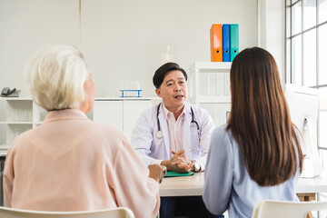 Obraz premium Asian male doctor consults with a senior female patient and her caregiver at a bright clinic office, offering medical advice and discussing treatment during a healthcare checkup session.