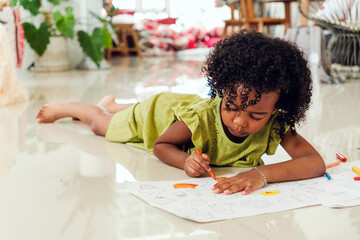 Cute Black girl in green dress lying on floor, coloring with crayons on large paper. Indoor...