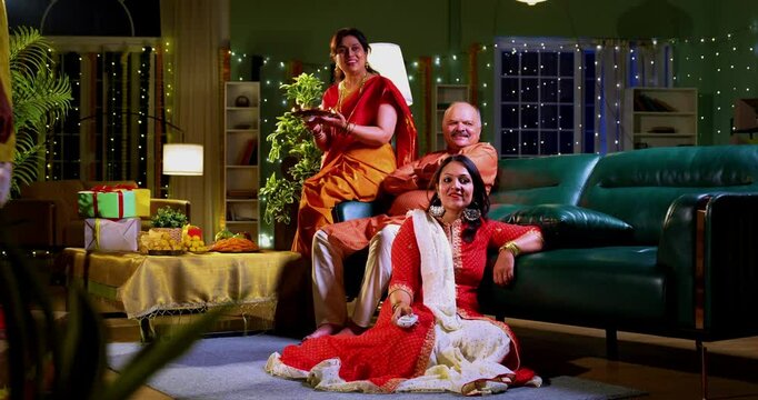 Son seeking Diwali blessings from parents, Indian family in festive home with senior mother in saree, father, and young daughter watching funny show on TV together after son greets with warm gesture