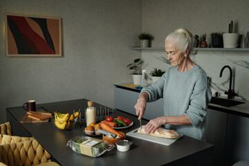 Senior woman slicing bread on a cutting board in contemporary kitchen filled with fresh ingredients like fruits, vegetables, and dairy products