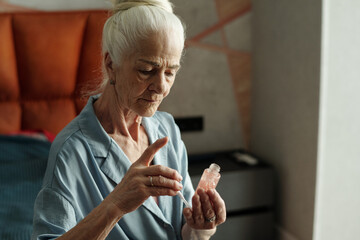 Elderly woman with white hair bun holding serum bottle while sitting indoors wearing a light blue shirt
