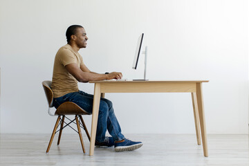 Side profile of African American man sitting at desk and working attentively on a desktop computer, young black freelancer guy browsing internet at home office, copy space