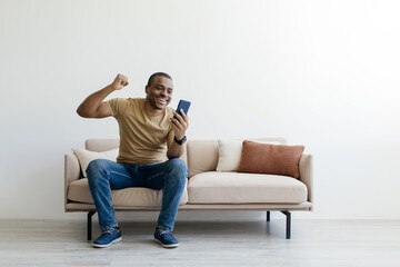 Great News. Joyful Black Young Man Looking At Smartphone Screen And Celebrating Success While Sitting On Couch At Home, African American Male Exclaiming With Excitement And Raising Fist