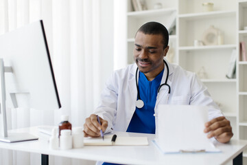 Young African American male doctor in white coat writing notes at desk, portrait of happy black...