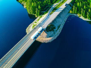 Aerial view of cargo truck and fuel tanker truck driving on asphalt bridge road over blue lakes water in Finland.