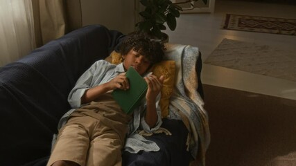 Young African American boy lying comfortably on cozy couch, reading green book as natural sunlight filling warm, inviting room