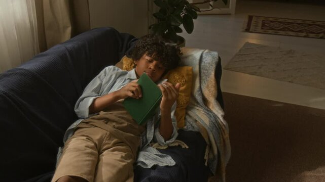 Young African American boy lying comfortably on cozy couch, reading green book as natural sunlight filling warm, inviting room