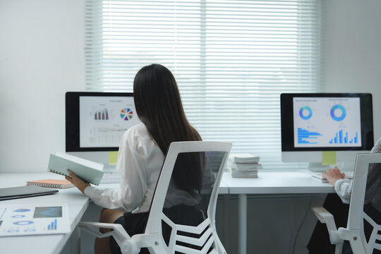 Two businesswomen are working on computers displaying charts and graphs, analyzing data and collaborating in a modern office environment with one of them holding a notebook