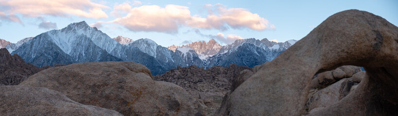 A person in red walks beneath Mobius Arch just after sunrise as the snowy Sierra Nevada ridge catches pink morning light, captured May 27, 2019