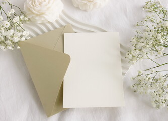 Blank paper card mockup, envelope, white flowers and white table decor candles flowers on white linen textured background. Copy space.Wedding invitation mockup, greeting card, holiday concept.