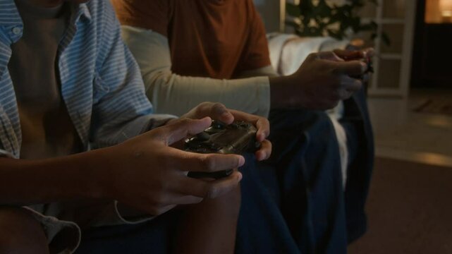 Closeup of unrecognizable father playfully interfering in gaming process as messing with controller of his son while playing together on couch in cozy, warm lit apartment