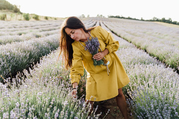 woman in yellow dress stands in lavender field, gently holding bouquet of lavender flowers. She appears serene and contemplative, enjoying beauty and fragrance of blossoms around her.