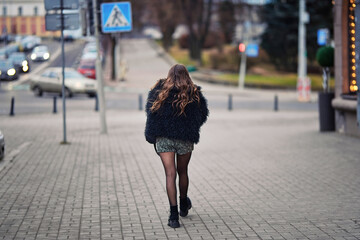 Stylish young woman in trendy outfit walking alone down quiet city street with moody atmosphere on cold overcast day. Rear view of woman in furry black coat, shorts and black tights walking alone