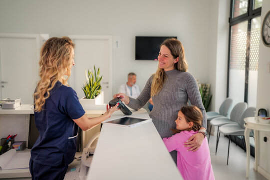 Pregnant woman paying with contactless payment at the clinic reception, while her daughter is hugging her, showing a modern healthcare payment process