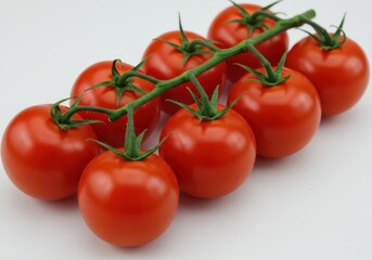 Cluster of fresh, ripe red cherry tomatoes still attached to the green vine, displayed on a white surface