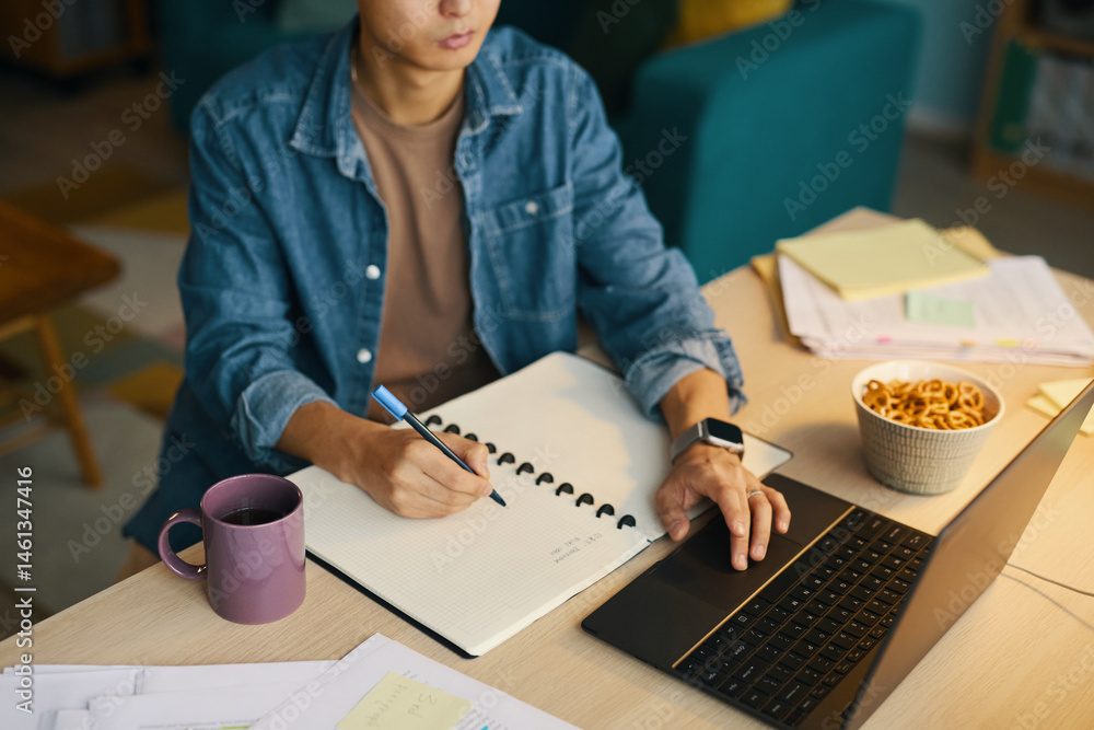 Poster male student writing notes on notepad while studying at desk with laptop open and various papers sca