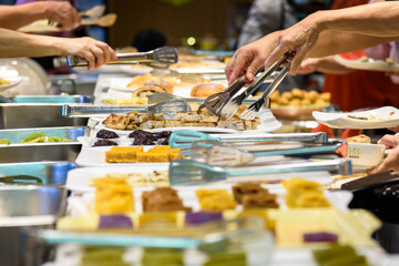 Hands with tongs select assorted dishes at a bustling buffet.