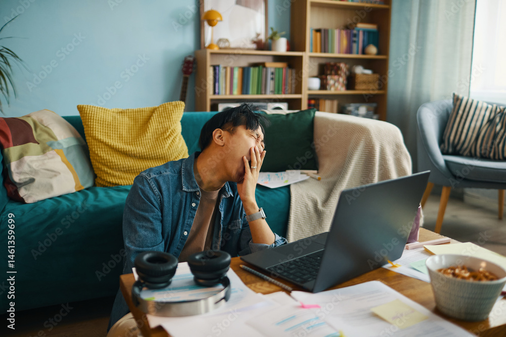 Poster man feeling exhausted while working on laptop in home office with bookshelves and couch background c