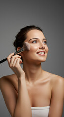 Fototapeta premium Radiant Beauty: Smiling Woman Applying Makeup Brush for Flawless Skin & Natural Glow - Cosmetics, Beauty, Skincare