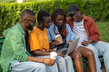 Interested group of african american students studying with laptop, looks webinar, sits outdoor on bench in park together. University classmates black friends discuss class project outside. 