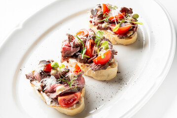 Three gourmet bruschetta slices with thinly sliced beef, cherry tomatoes, microgreens, grated cheese, and sauce drizzle, served on a white plate. A fresh and elegant appetizer.