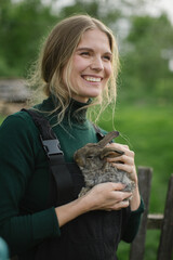 A happy blonde woman holds a rabbit in her arms. Aesthetics of rural life.