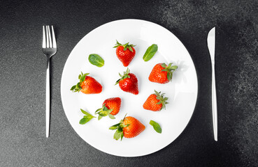 Fresh strawberries arranged on a white plate with mint leaves beside cutlery