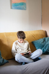 Fototapeta premium Young boy sitting on sofa playing with smartphone at home