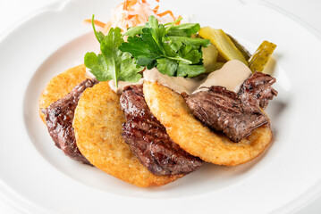 Grilled beef steak with crispy potato pancakes, garnished with fresh parsley, pickles, and creamy sauce on a white plate. Served with shredded carrots and cabbage. Delicious and appetizing meal.