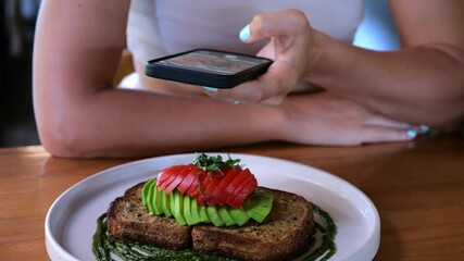 Young woman capturing artisan avocado toast with tomato and pesto, documenting culinary moment using smartphone while dining in trendy restaurant setting - Powered by Adobe