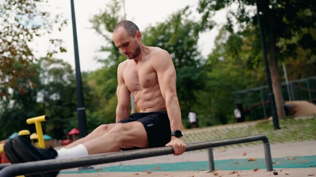A muscular man performs dips on parallel bars in a park. He focuses on his form and strength as he completes each repetition. The park setting provides a natural backdrop for this fitness-focused shot