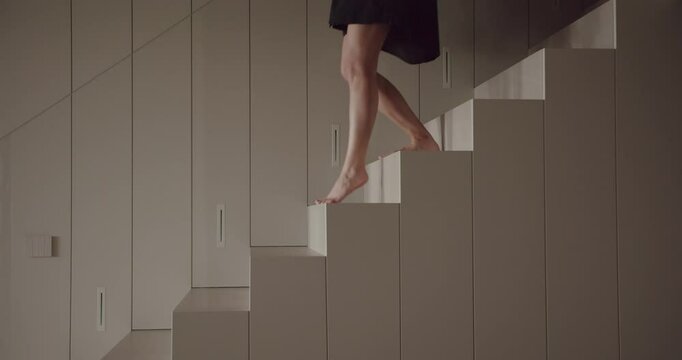 A woman in a black dress walks elegantly on a sequence of high, neutral-toned geometrically shaped cabinets in a modern, minimalist room. The image captures a sense of balance and minimalism.