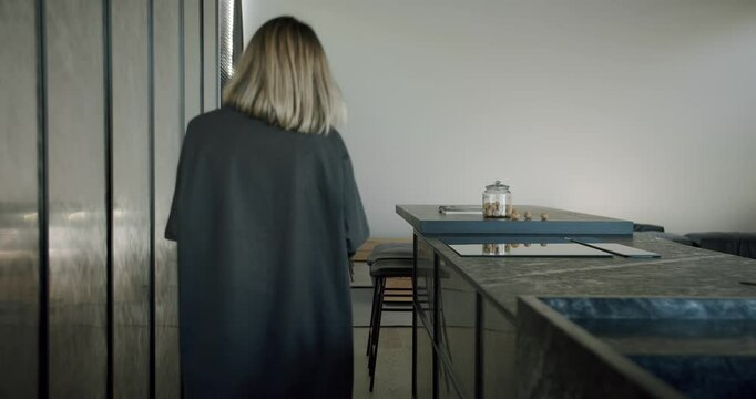 A young woman in a stylish black robe walks away in a sleek, modern kitchen. The scene captures a tranquil, domestic atmosphere with minimalist appeal. Modern Kitchen with Young Woman