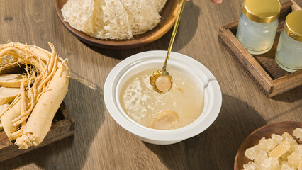 A white porcelain bowl of bird's nest is placed on a dark brown wooden table, with a slice of ginseng on a spoon. Other ingredients such as rock sugar, ginseng are placed on wooden plates.