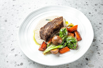 A gourmet dish featuring a juicy beef steak topped with a creamy sauce, served with roasted vegetables including carrots, potatoes, and greens, elegantly plated on a white plate against a textured cou