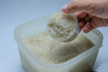 A human hand holding a plastic cup filled with raw white rice above a plastic container full of rice. Suitable for use in articles, advertisements, or educational materials about food ingredients