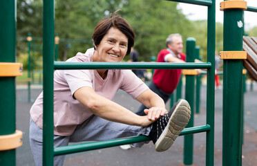 Obraz premium Elderly woman doing stretching exercises on the parallel bars at an outdoor sports ground