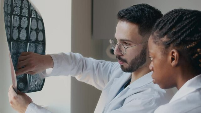Side view of male Middle Eastern neurologist discussing with female colleague patients treatment plan looking at MRI scan results while cooperating at workplace