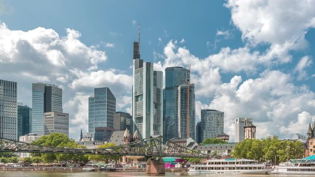 Hyperlapse of the historical Eiserner Steg (Iron Bridge) over the Main River in Frankfurt, Germany. Skyscrapers skyline and historic buildings reflect in the water under a cloud-filled sky timelapse
