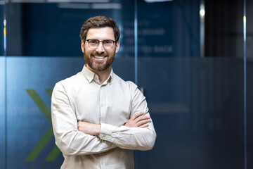 Portrait of a young businessman in a shirt and glasses standing in an office, arms crossed on his...