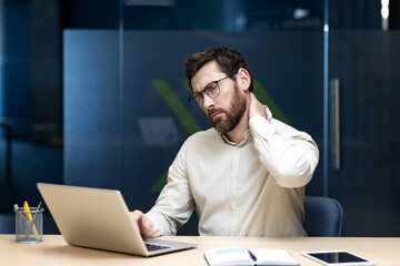 A tired young man sits in the office at his desk, rubbing his neck with his hand, feeling a dull...