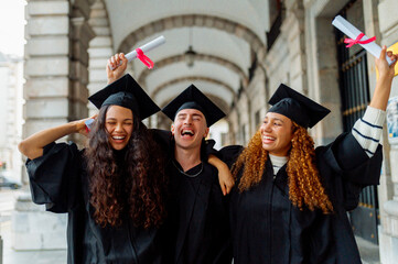 Group of cheerful multi ethnic graduates wearing gowns and mortarboards, holding diplomas and...
