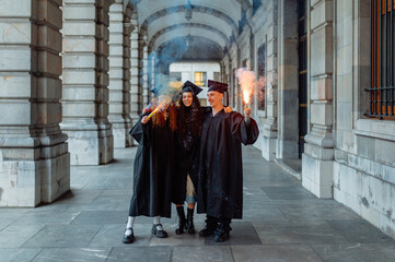 Three cheerful multiracial students celebrating their graduation day with smoke bombs in a...