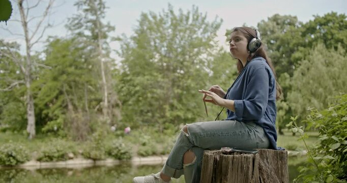 Young woman enjoys listening to music in nature. She sits on a tree stump by the lake and conducts the music with a twig, wearing headphones and casual clothes.