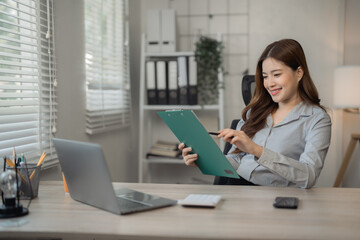 Smiling asian businesswoman pointing at clipboard and reviewing documents while working on laptop in modern office, business and technology concept