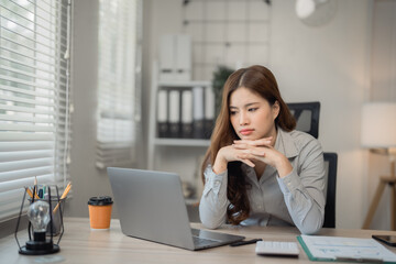 Obraz premium Young asian businesswoman working with laptop looking concerned while sitting at her desk in modern office with documents, calculator, coffee and light bulb