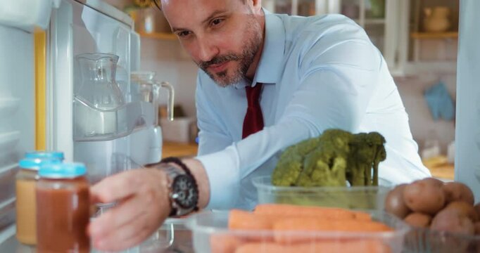 A businessman opens the refrigerator and takes some porridge for the baby.