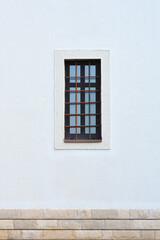 A small, dark window with multiple panes and metal bars is set in a textured white wall above a stone foundation