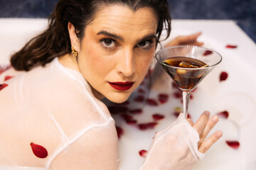 Elegant woman in milk bath holding cocktail surrounded by red petals