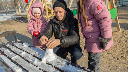 Grandfather with granddaughters making a little snowman in sunny winter day